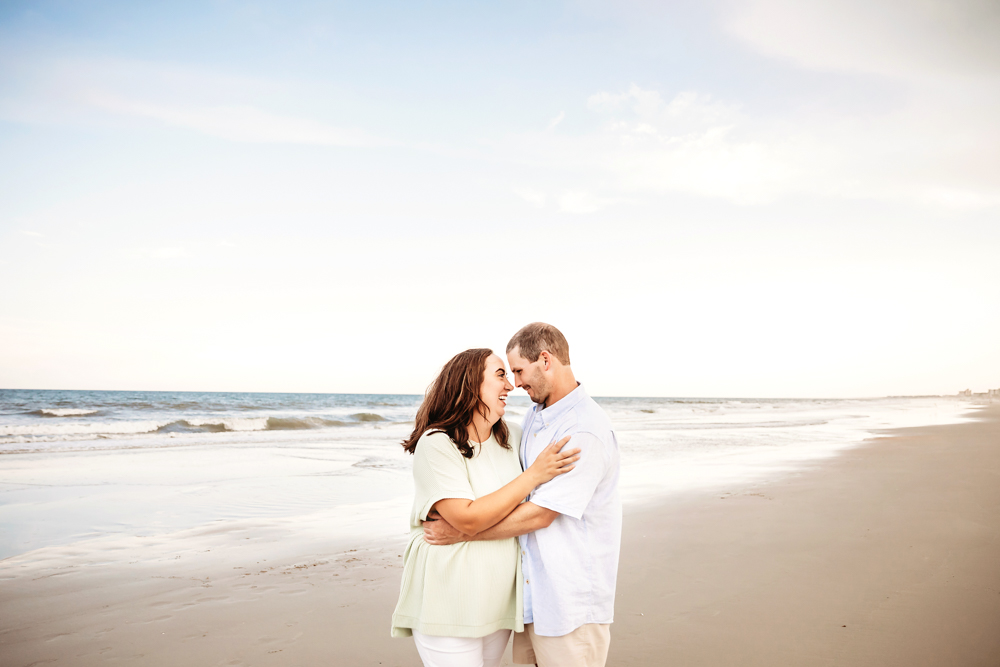 Mom and dad embrace, smiling softly at each other with the ocean waves behind them.