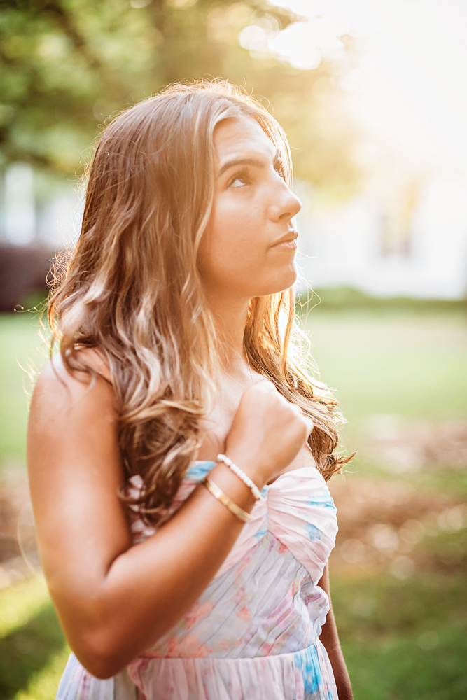 Senior session photo taken in open field at golden hour, showing a looking-away pose in floral dress from a full-body angle