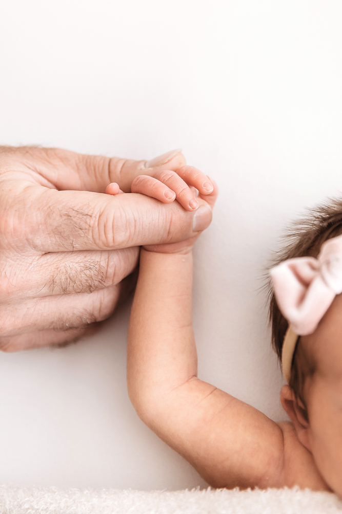 Close-up of a newborns tiny hand holding fathers thumb beside her head with a bow headband.