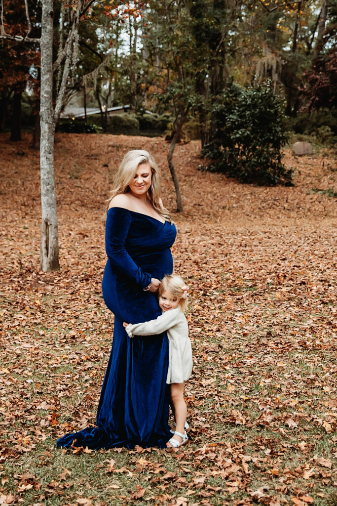 Young daughter hugging pregnant mom in blue velvet dress during fall session with golden leaves in Florence SC