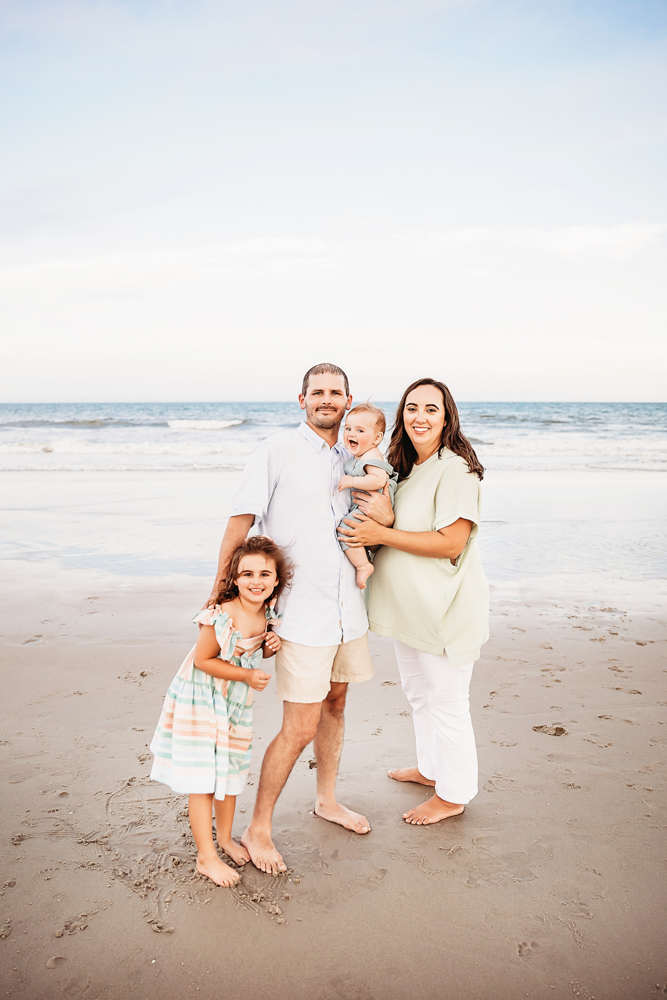 The full family of four poses happily at the beach, all smiling with the sea and sky behind them.