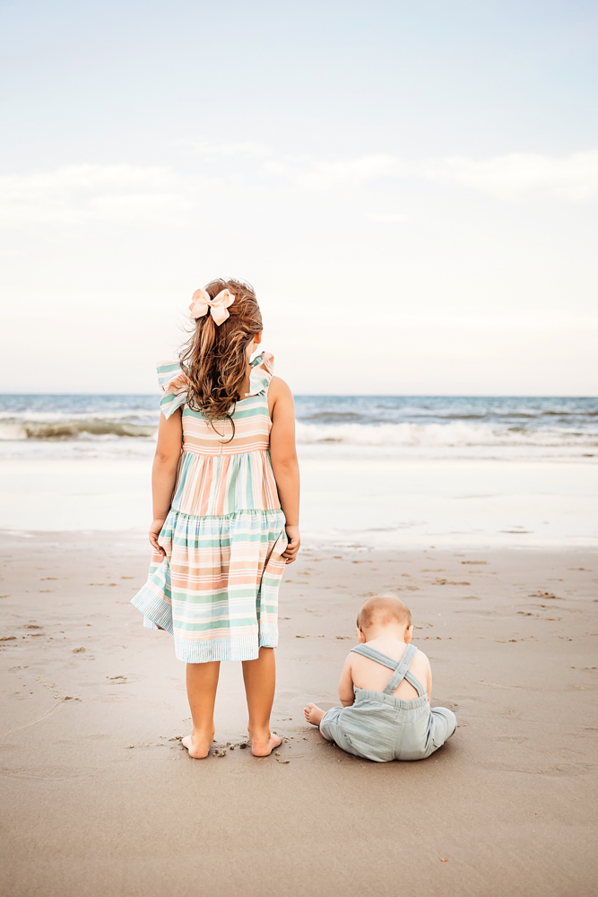Big sister stands tall beside her baby brother who is sitting in the sand, both facing the ocean.