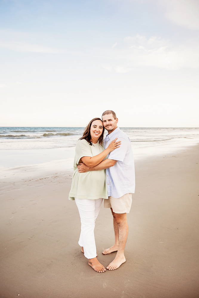 Mom and dad stand together hugging, smiling gently with their heads touching and the beach stretching out behind them.