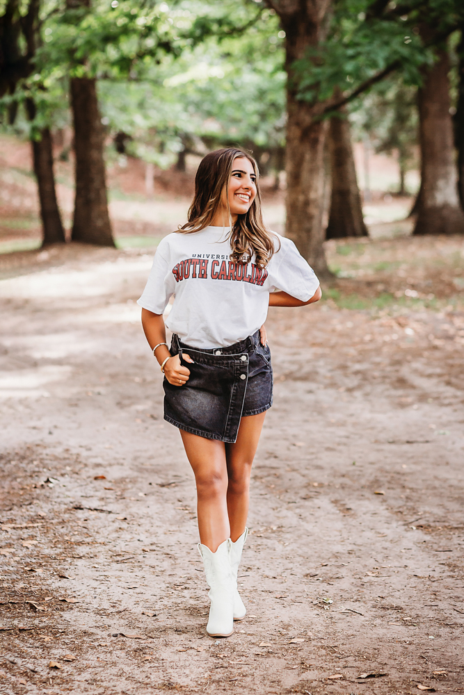 Senior session photo taken in gravel path at golden hour, showing a standing pose in USC shirt and denim skirt from a close-up angle