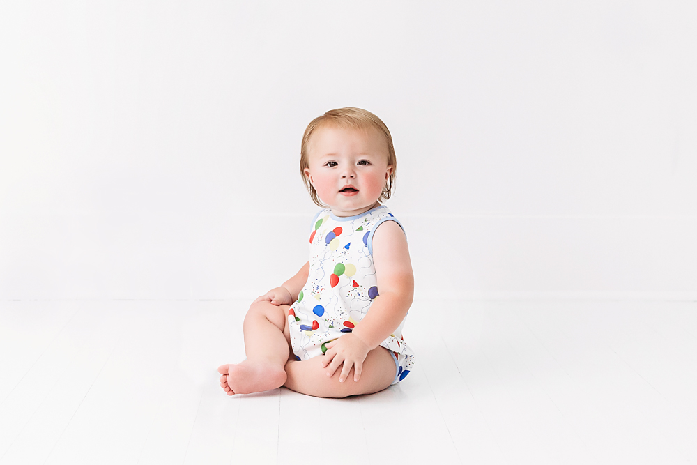 Levi sitting upright on a white floor wearing a balloon-print romper, calmly posing for his first birthday portraits at Reflection Images by Tracy Rowell in Florence SC