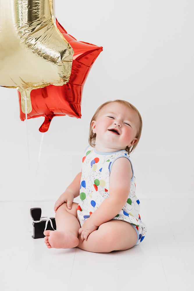 Levi laughing with joy next to a gold and red balloon bouquet, wearing a festive birthday romper on a bright white studio floor at Reflection Images by Tracy Rowell in Florence SC