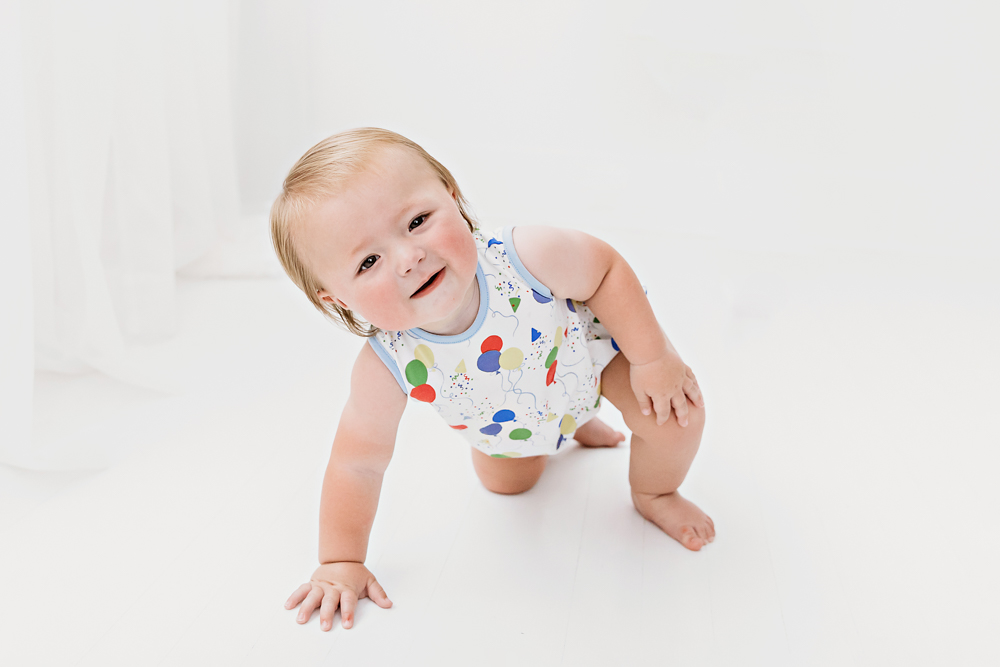 Levi mid-crawl with a happy expression, wearing a balloon-themed romper in a white studio setup with natural light at Reflection Images by Tracy Rowell in Florence SC