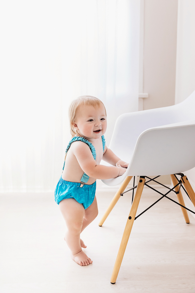 Levi standing beside a modern white chair with soft natural light and blue romper, smiling proudly during his milestone session at Reflection Images by Tracy Rowell in Florence SC