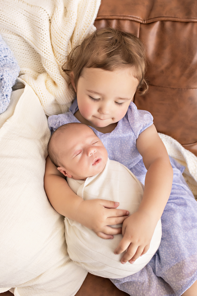 big sister cuddling swaddled newborn on leather couch with knit throws Florence SC sibling photos