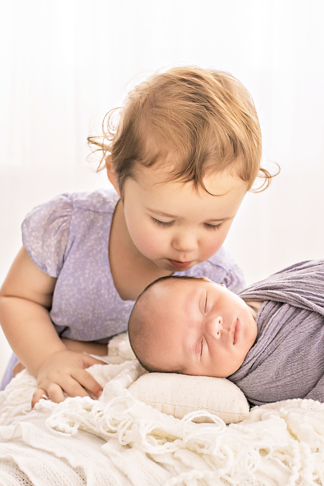backlit moment sister leaning to kiss swaddled newborn on white bed Reflection Images Florence SC professional photography studio
