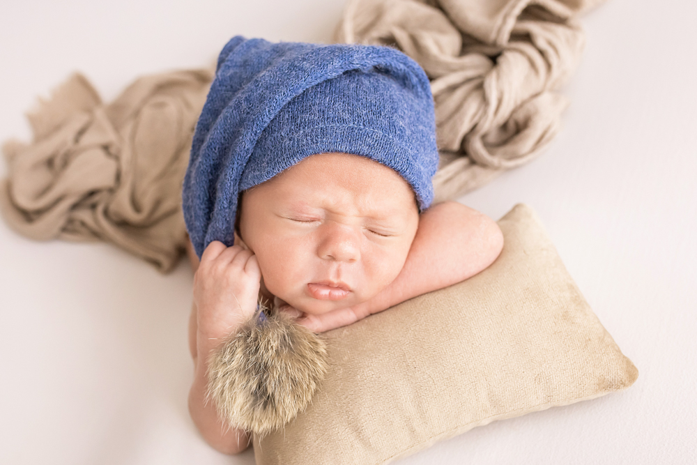 newborn with blue sleepy cap and fur pom resting on tan pillow neutral layers Florence SC Reflection Images