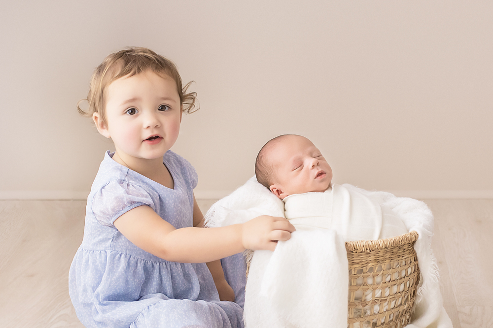 toddler sister seated beside basket touching newborn swaddle neutral studio Florence SC