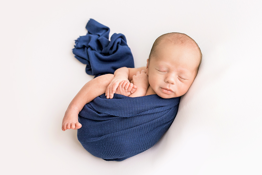 newborn Walker snug in navy wrap with crossed arms peeking out on white blanket Florence SC Tracy Rowell