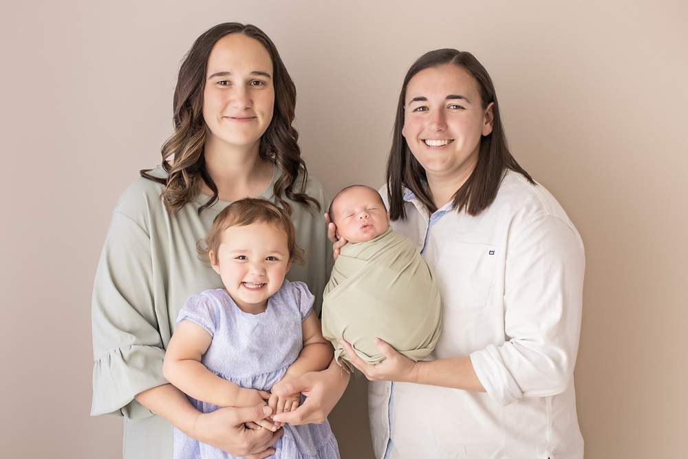 two moms and daughters studio portrait holding newborn in sage wrap against beige wall Florence SC