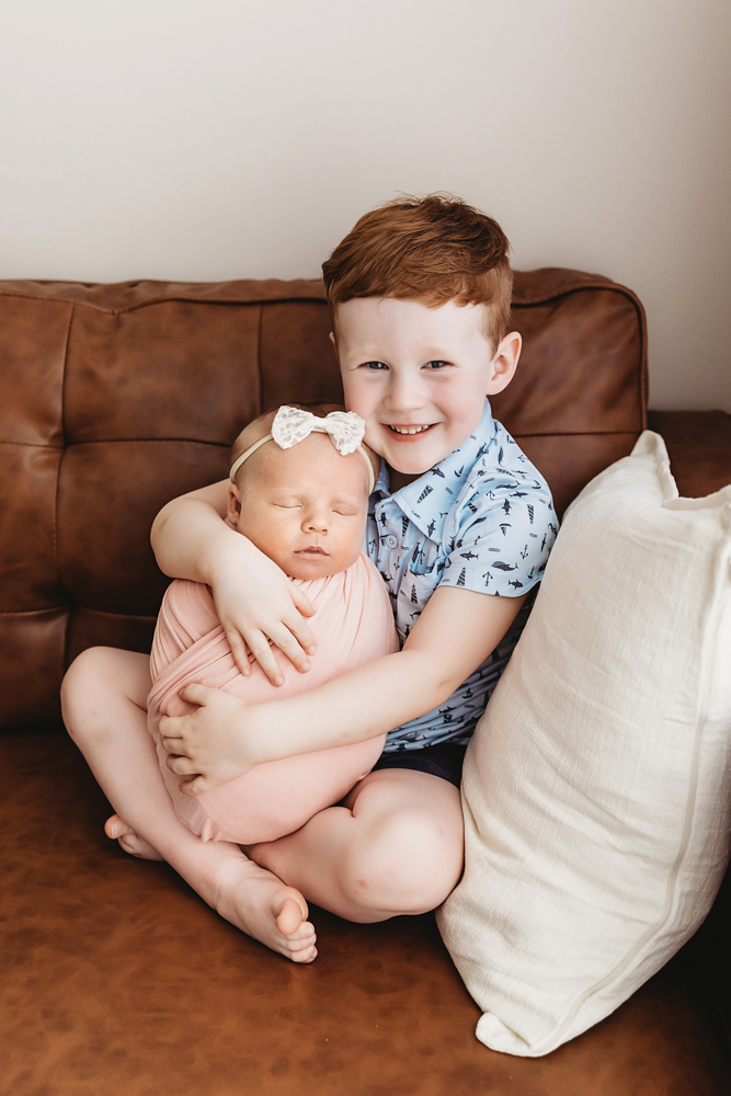 Big brother holding Millie Jo on a brown leather couch, both calm and cozy at Reflection Images by Tracy Rowell in Florence SC