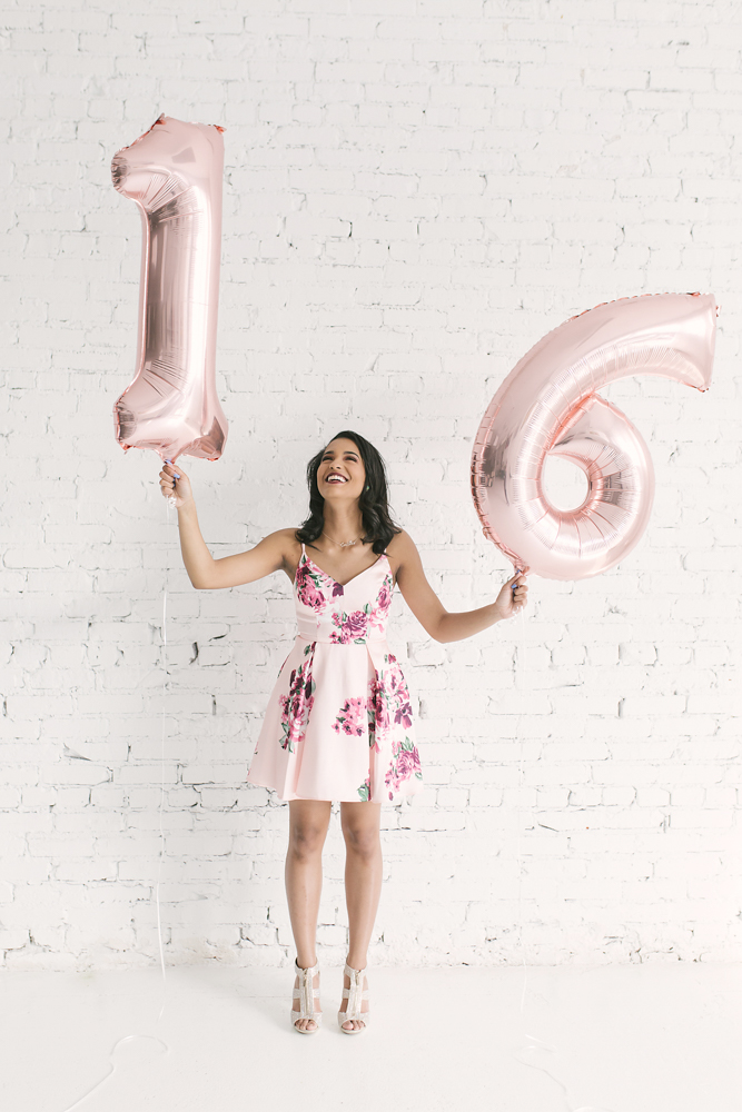 Connie standing against a white brick wall holding metallic rose gold '16' balloons, wearing a pink floral dress and heels at Reflection Images by Tracy Rowell in Florence SC