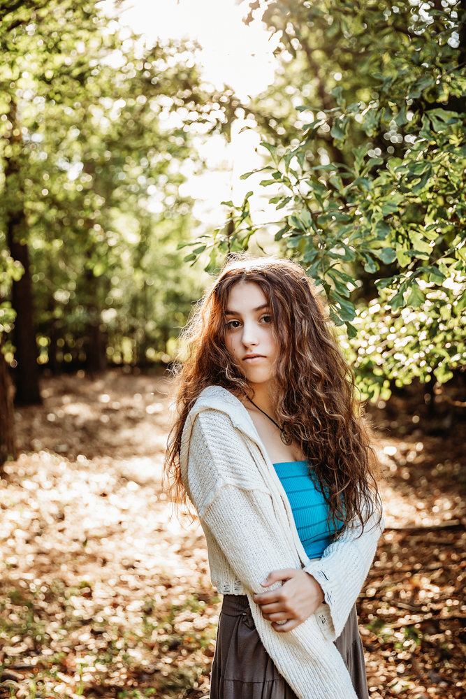Eve with long wavy hair, turquoise bandeau and cream sweater, soft expression against leafy forest background…