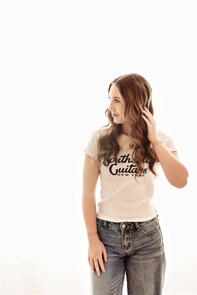 Addy standing in soft window light, gently touching her hair with loose waves, wearing a Southside Guitars tee and jeans at Reflection Images by Tracy Rowell in Florence SC