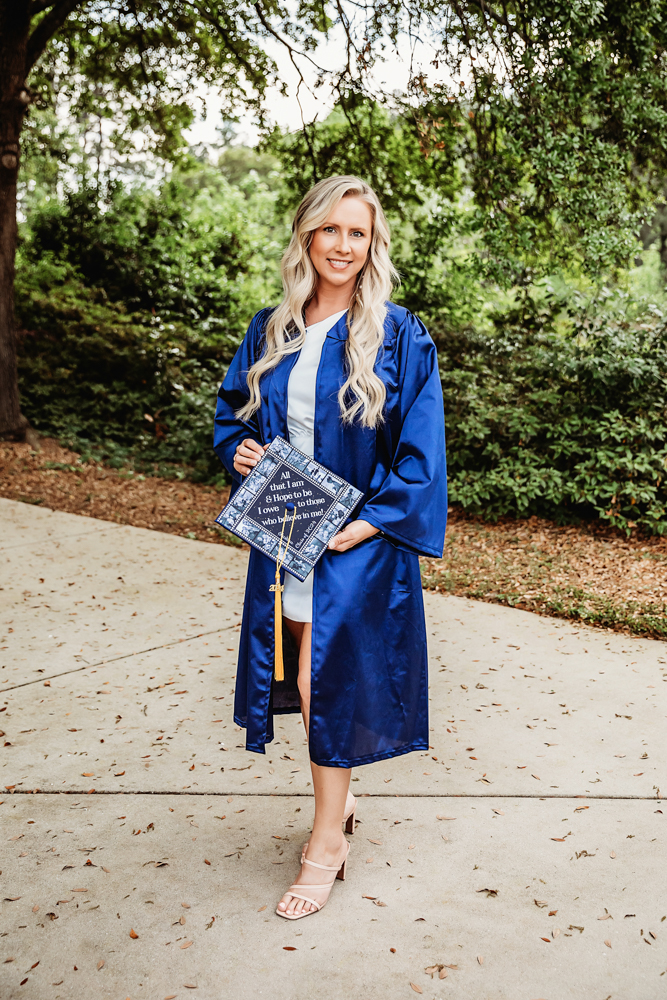 Kristina in a navy blue graduation gown holding her decorated cap while smiling on a sidewalk surrounded by greenery at Reflection Images by Tracy Rowell in Florence SC