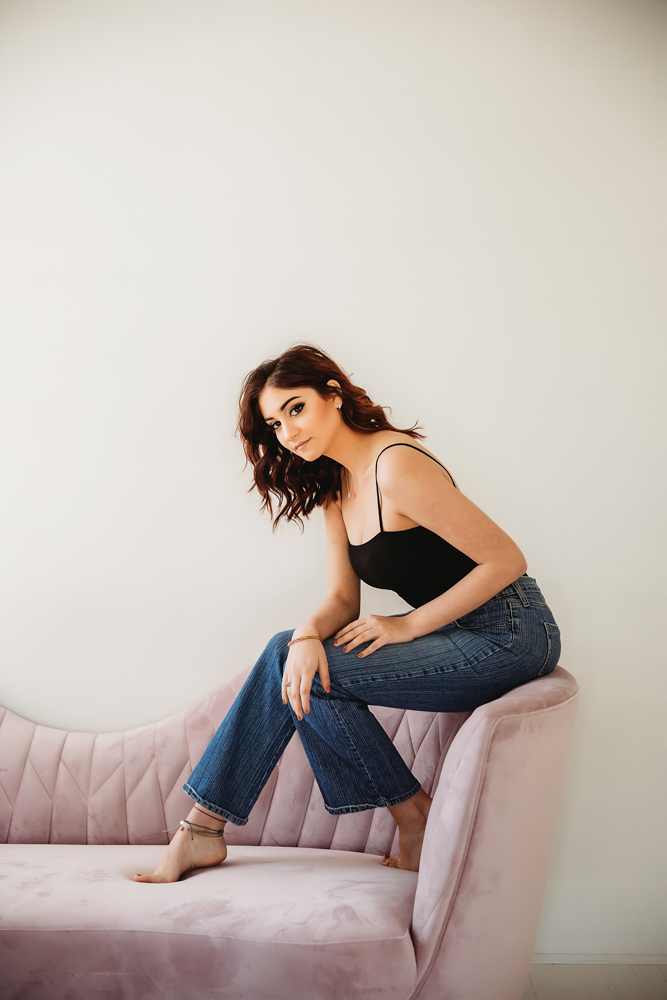 Patricia sitting barefoot on blush pink sofa wearing black top and jeans at Reflection Images by Tracy Rowell in Florence SC