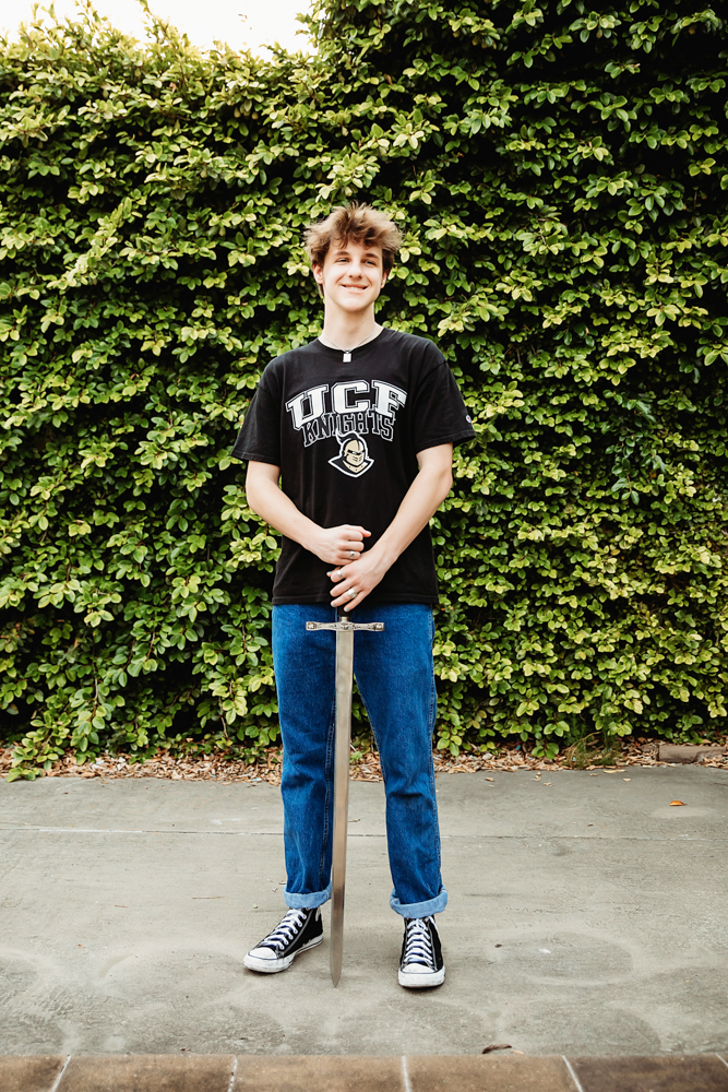 Jack standing with a medieval-style sword, wearing a UCF Knights shirt and jeans in front of a lush green ivy wall at Reflection Images by Tracy Rowell in Florence SC