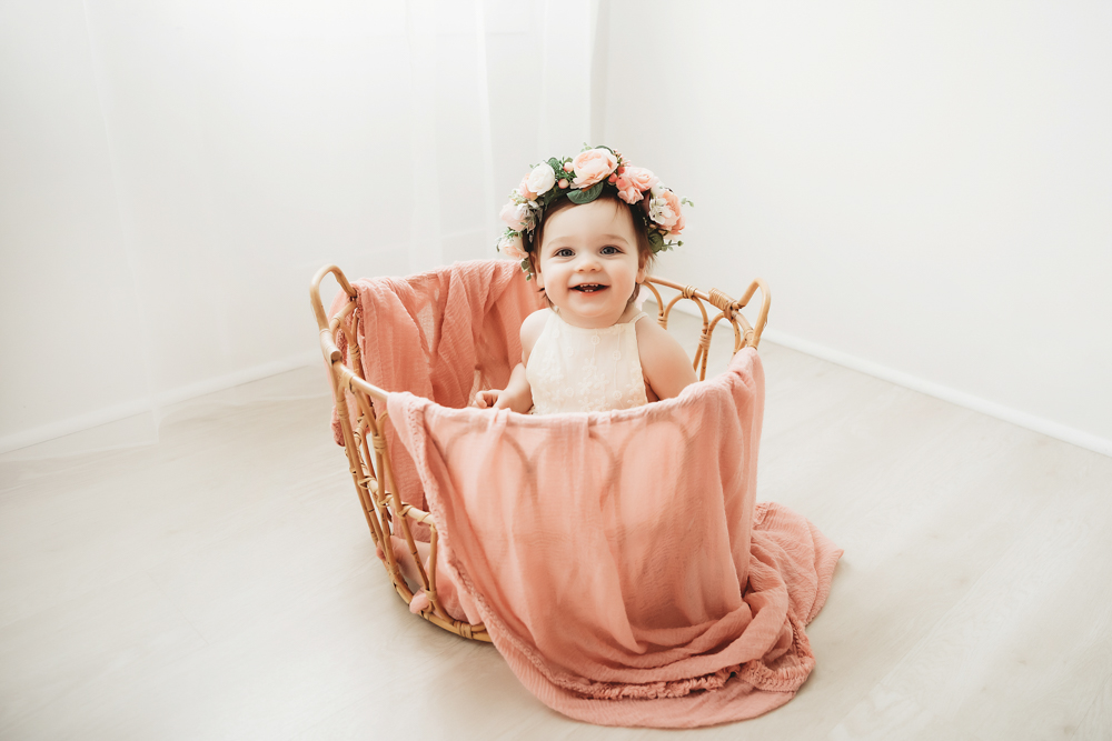 Aubrey in a floral crown and white romper smiling from inside a wicker basket with blush wrap at Reflection Images by Tracy Rowell in Florence SC