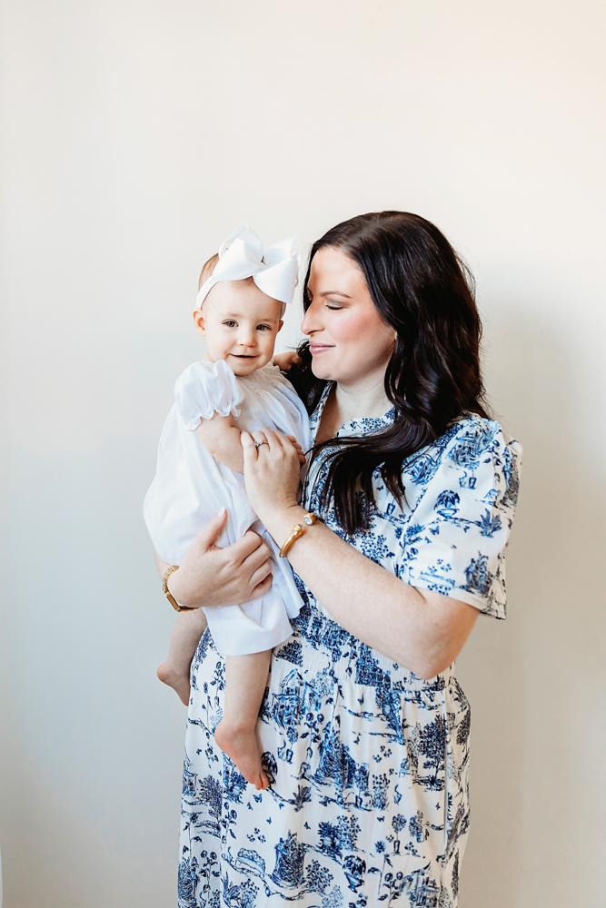 Mom holding Elsie in a white dress and bow, both smiling, studio neutral backdrop at Reflection Images by Tracy Rowell in Florence SC