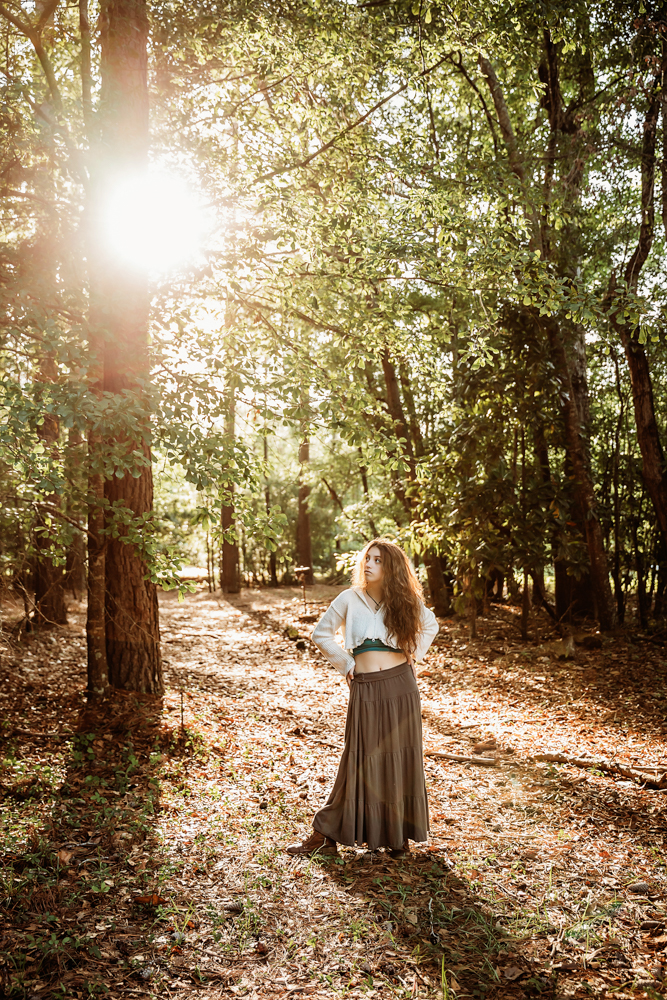 Eve in turquoise top and brown skirt, standing with hands on hips in golden sunlight among tall trees…