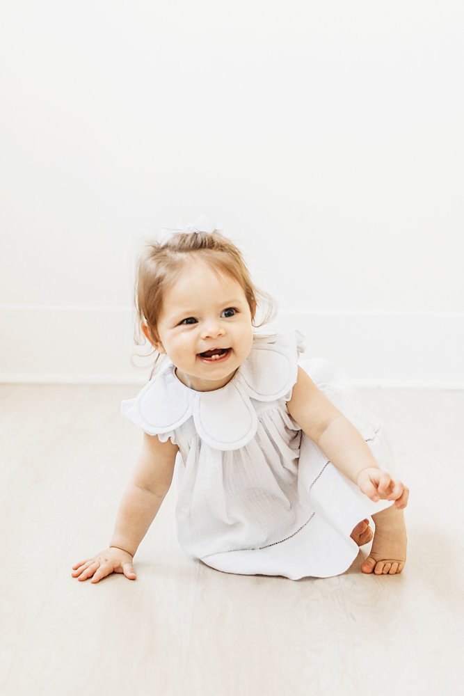 Baby girl crawling on light floor in white dress with oversized collar.