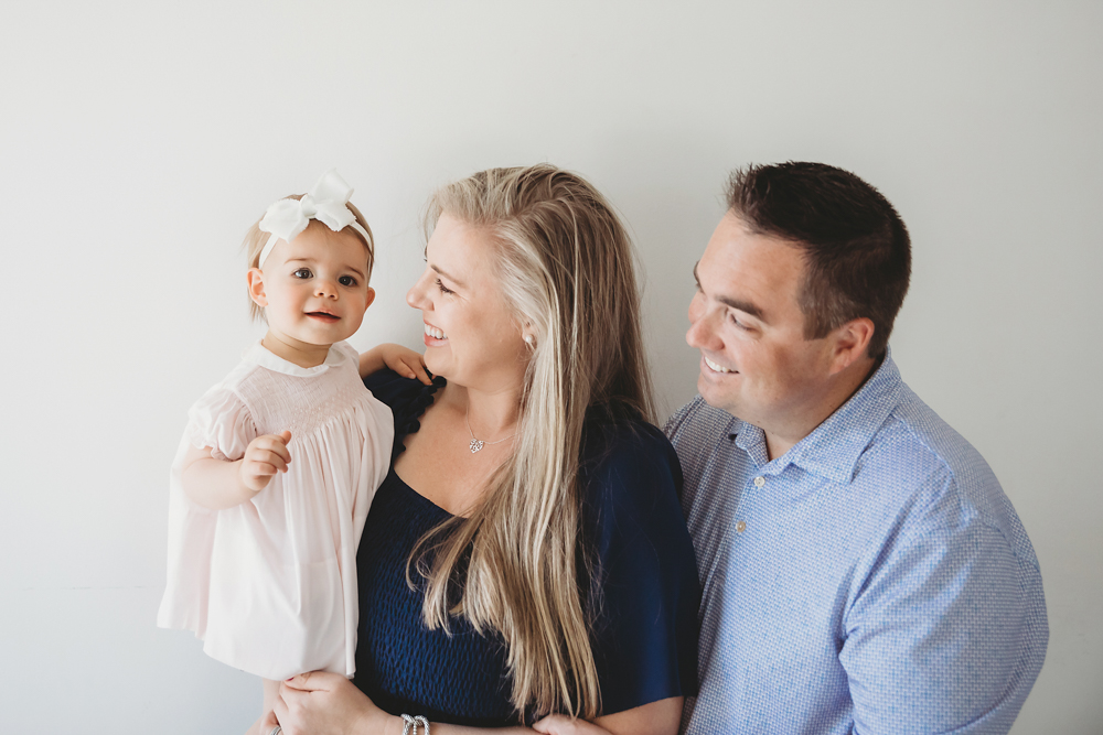 Grace held by both parents, smiling with her white bow and light dress, captured in soft natural light at Reflection Images by Tracy Rowell in Florence SC