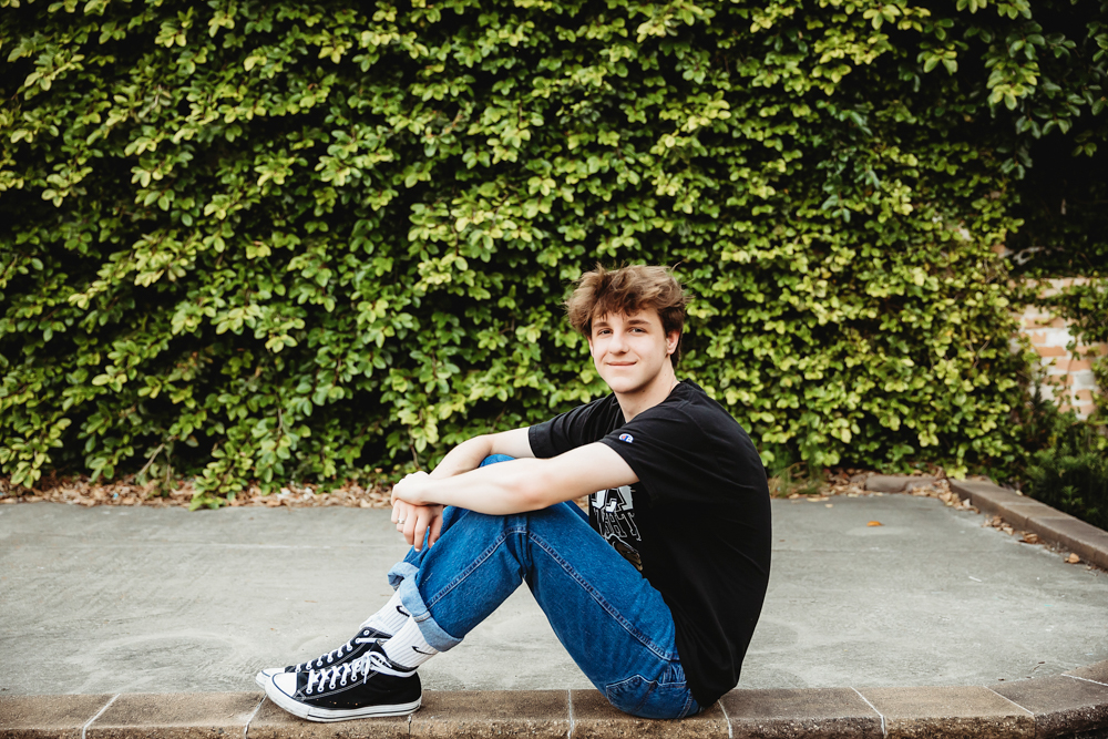 Jack seated casually in cuffed jeans and Converse sneakers against a deep green ivy backdrop during his downtown Florence senior session with Reflection Images by Tracy Rowell