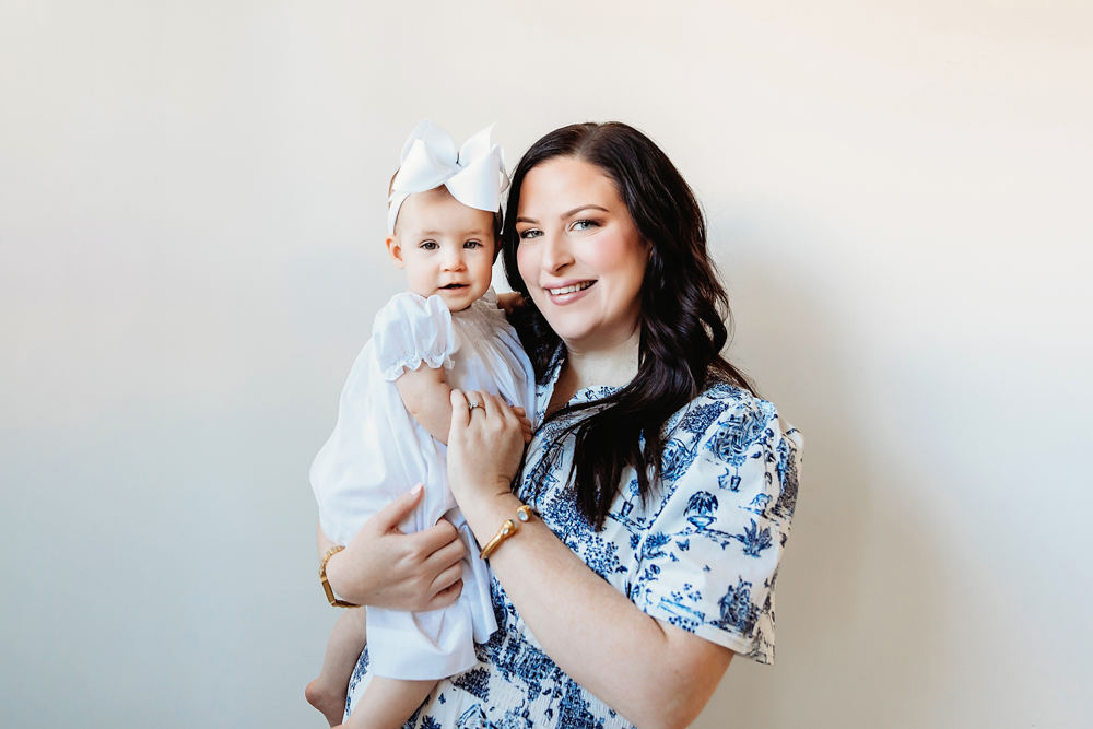 Mom holding Elsie in a white dress and bow, both smiling, studio neutral backdrop at Reflection Images by Tracy Rowell in Florence SC