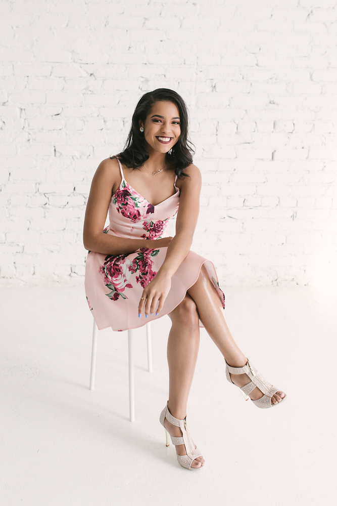 Connie seated on a white stool in her pink floral dress and glitter heels, smiling brightly with a white brick backdrop at Reflection Images by Tracy Rowell in Florence SC