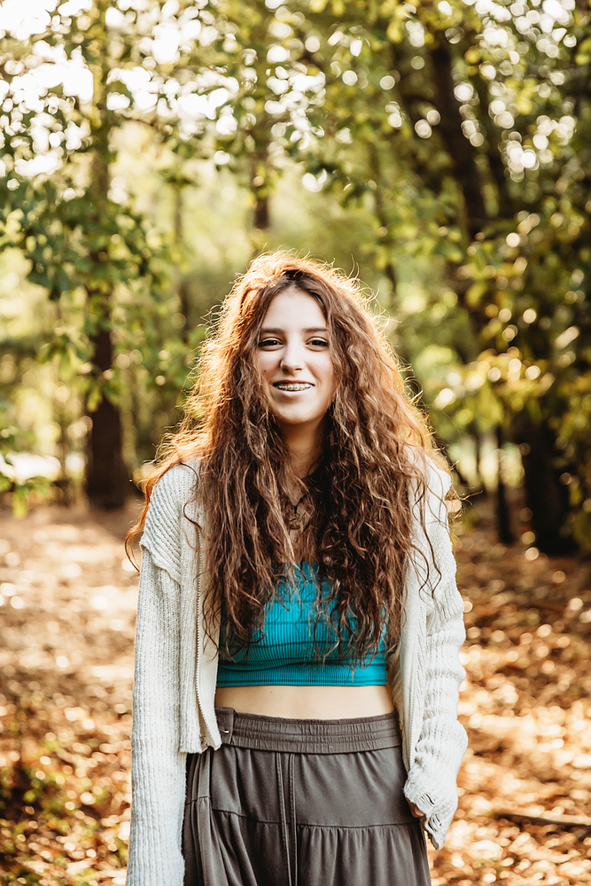 Eve with curly hair smiling brightly in turquoise top and cream sweater, warm golden forest light…