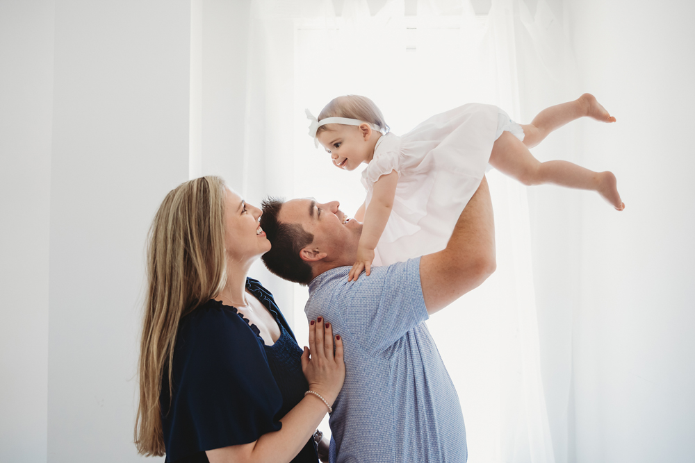 Grace being lifted above by dad with both parents smiling below, light streaming in from a sheer white-curtained window at Reflection Images by Tracy Rowell in Florence SC