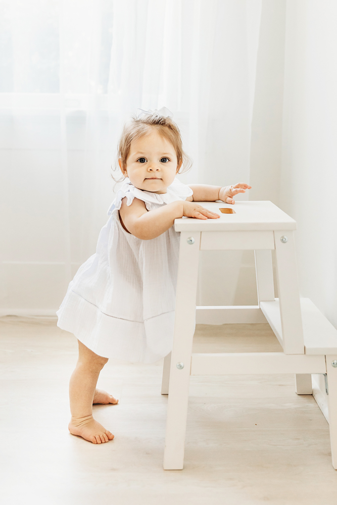 Birthday girl standing holding a white stepstool with sheer curtains in background.