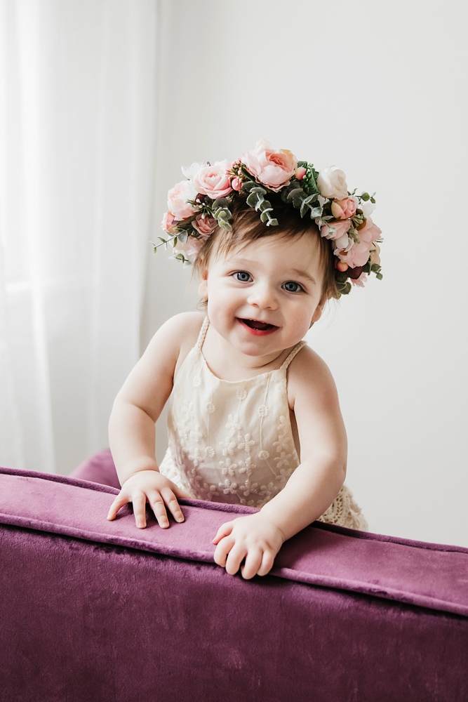 Aubrey standing at a velvet chair in a lace romper and floral crown, smiling with soft window light at Reflection Images by Tracy Rowell in Florence SC