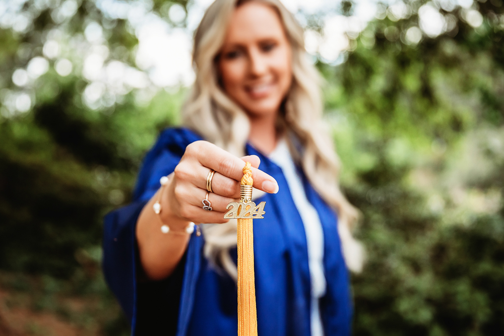 Macro shot of Kristina holding her gold 2024 tassel forward, with a soft blur of her gown and greenery behind at Reflection Images by Tracy Rowell in Florence SC