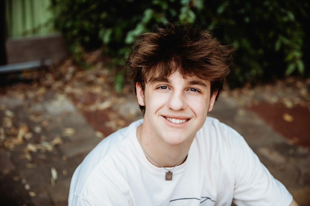 Close-up of Jack smiling with tousled hair, sitting outdoors on a brick path surrounded by foliage at Reflection Images by Tracy Rowell in Florence SC