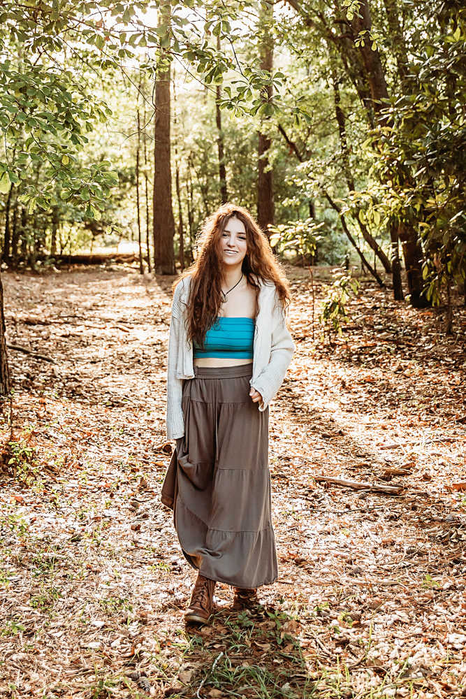 Eve walking on wooded path in turquoise bandeau, cream sweater, and long skirt, natural light…