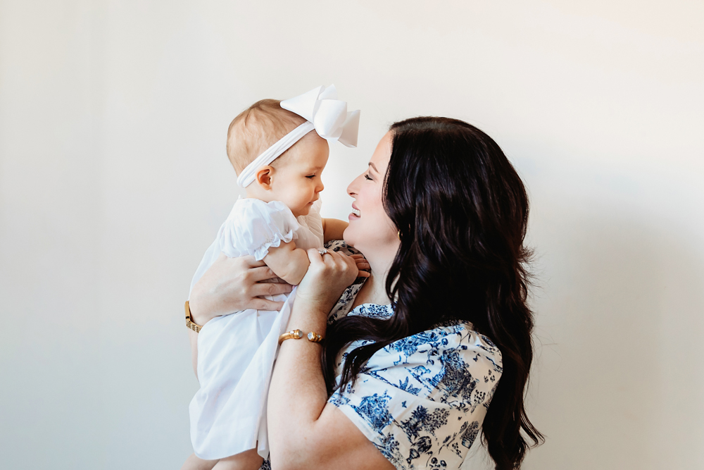 Mom holding Elsie in a white dress and bow, both smiling, studio neutral backdrop at Reflection Images by Tracy Rowell in Florence SC