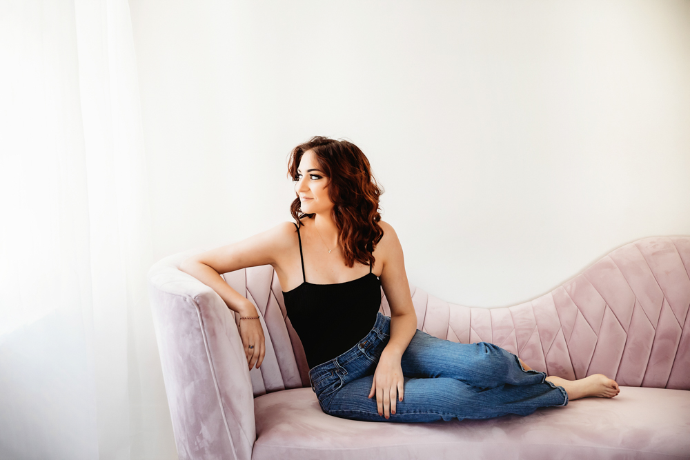 Patricia seated on pink sofa gazing out window light in black top and denim jeans at Reflection Images by Tracy Rowell in Florence SC