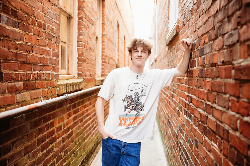 Jack leaning confidently in a narrow brick alley wearing a western tee and jeans during his senior session with Reflection Images by Tracy Rowell in Florence SC