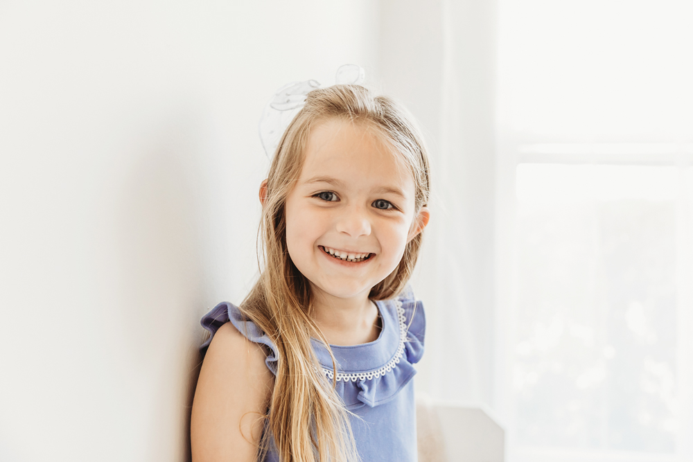 Older sister standing against wall smiling, sunlight coming through window.