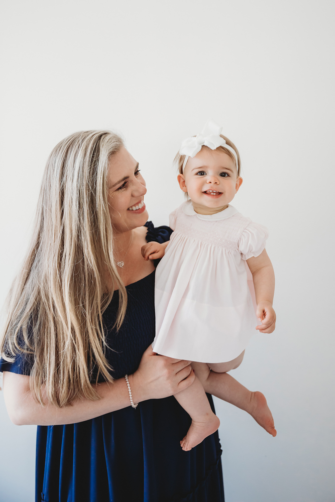 Grace in moms arms, smiling brightly in her white dress and bow with a clean studio backdrop at Reflection Images by Tracy Rowell in Florence SC