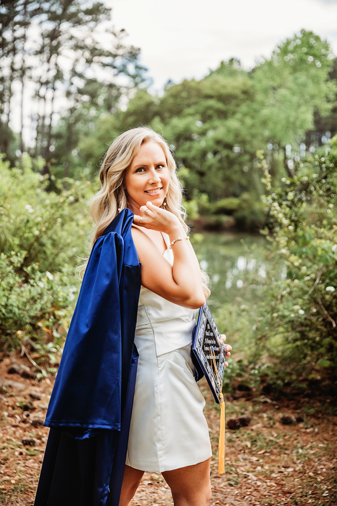 Kristina smiling with her cap over her shoulder, white dress peeking through, among pine needles and soft light at Reflection Images by Tracy Rowell in Florence SC