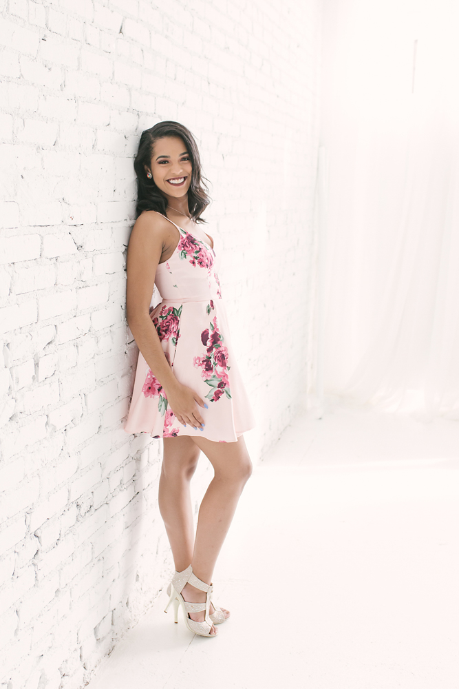 Connie leaning against a white brick wall in her floral dress, smiling confidently in soft natural light at Reflection Images by Tracy Rowell in Florence SC