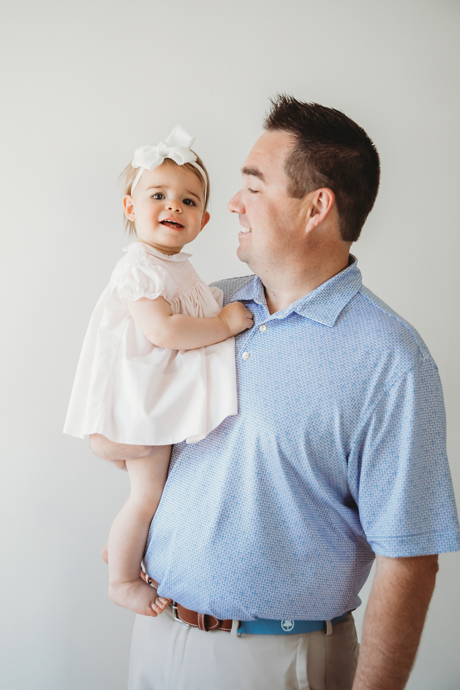Grace in a white smocked dress with a big white bow, smiling while being held by dad against a light background at Reflection Images by Tracy Rowell in Florence SC
