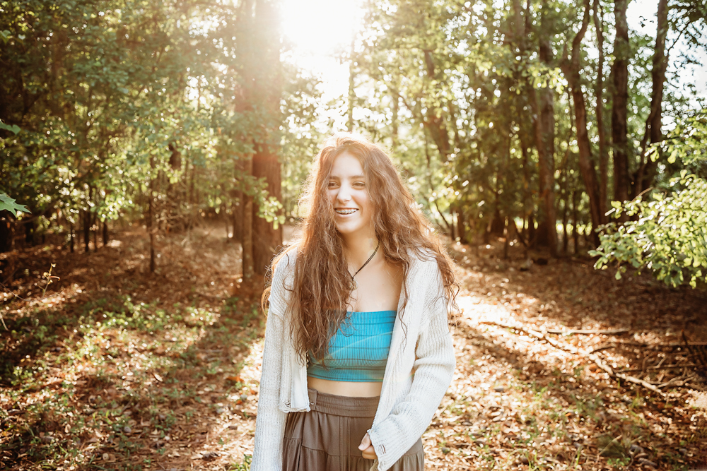 Eve smiling with long wavy hair, turquoise top, cream sweater, in forest sunlight…