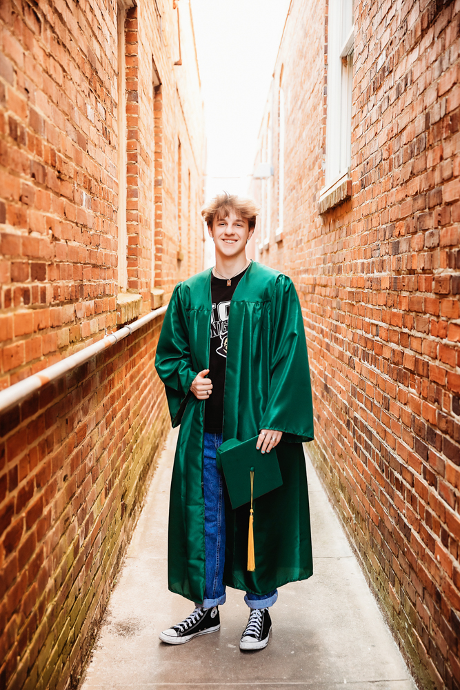 Jack smiling in a green graduation cap and gown, posed in a brick alleyway downtown Florence for his senior photos with Reflection Images by Tracy Rowell